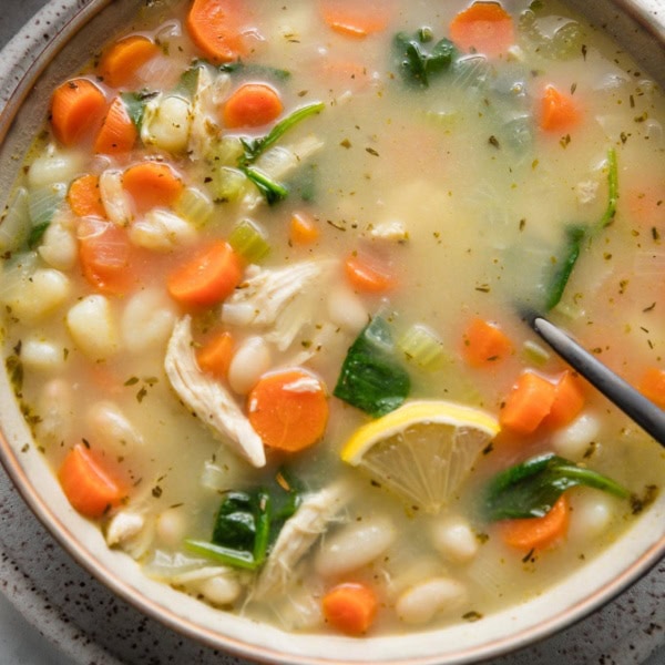 Close up of a bowl of white bean soup with veggies, mini gnocchi, and shredded chicken, with fresh parsley and a lemon wedge sprinkled on top.