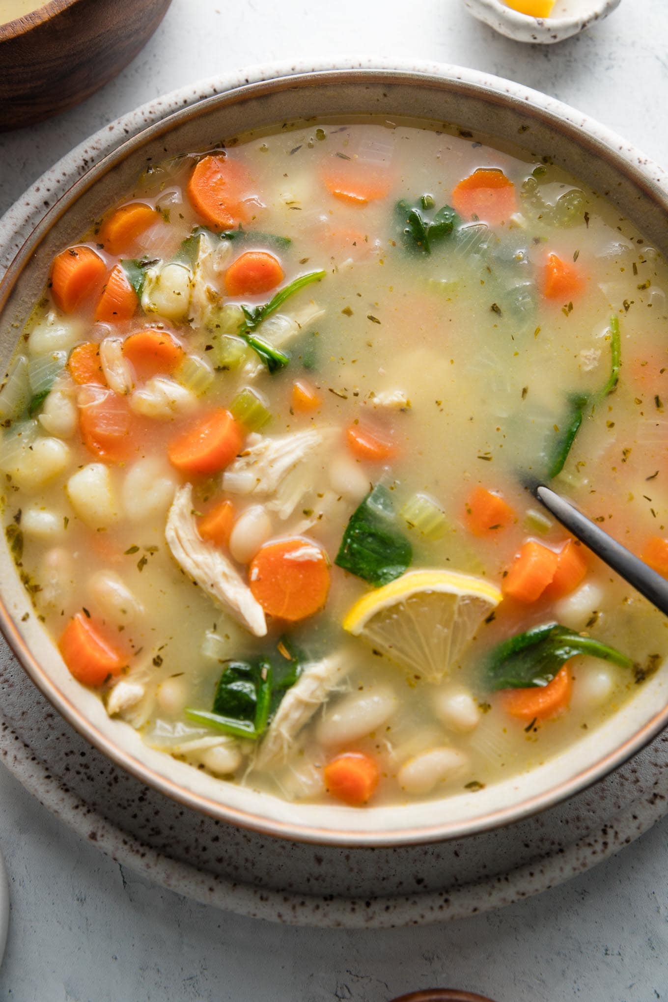 Close up of a bowl of white bean soup with veggies, mini gnocchi, and shredded chicken, with fresh parsley and a lemon wedge sprinkled on top.