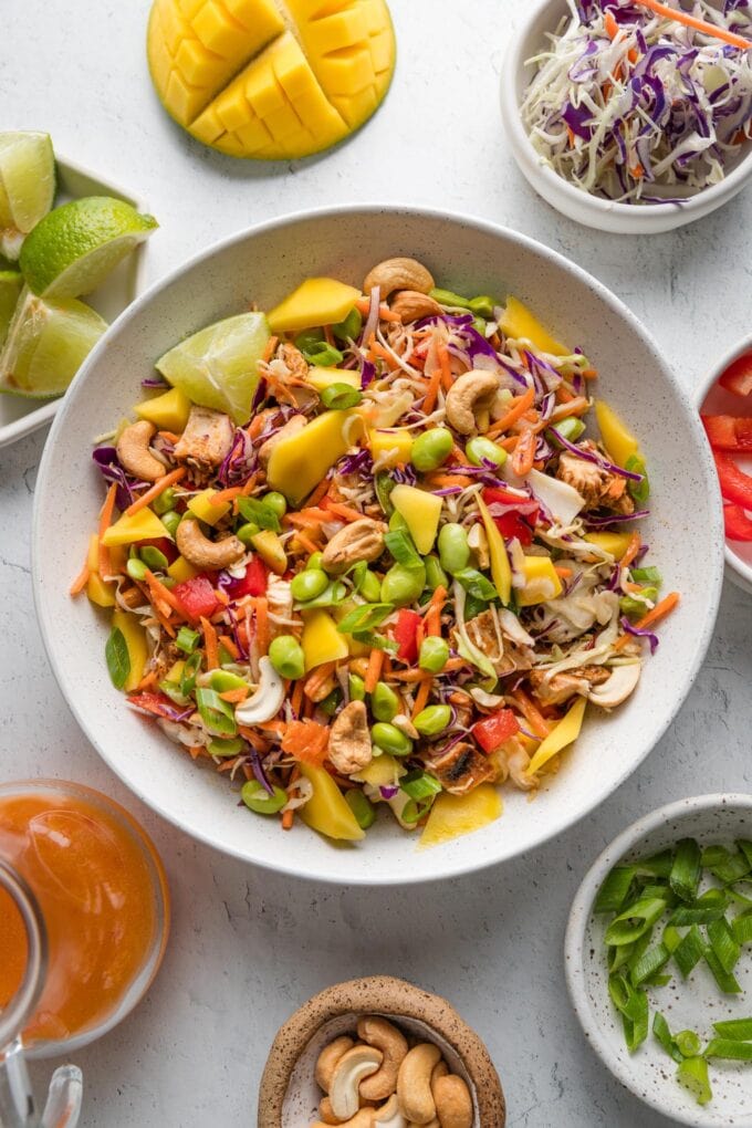 Overhead view of a chopped Thai-inspired chicken salad with mango, cashews, cabbage, green onions, and extra sweet chili dressing scattered on the counter nearby.