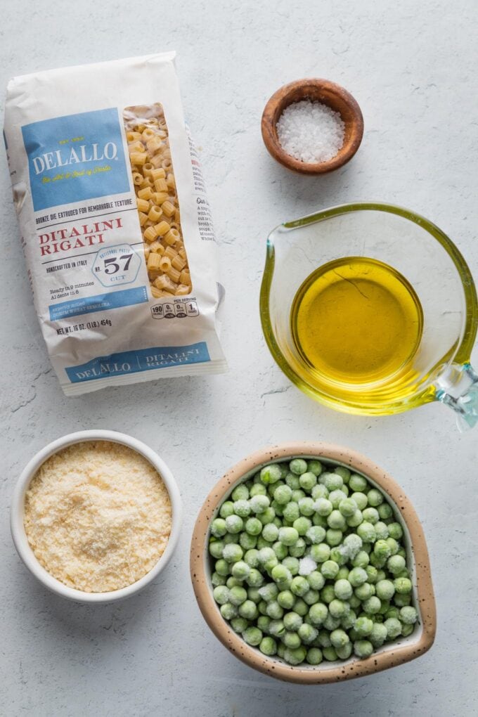 Overhead image of a package of ditalini, olive oil in a liquid measuring cup, peas, Parmesan, and salt.
