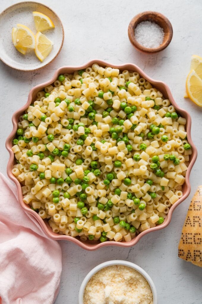 Countertop set with a pasta dish of ditalini, peas, and Parmesan, plus extra garnishes nearby - lemon edges, salt, pepper, and more Parm.