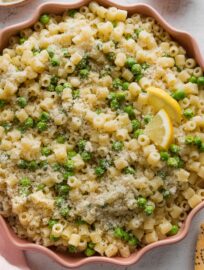 Close up of ditalini with peas, Parmesan, and black pepper served in a pink scalloped bowl with extra Parmesan on the table nearby.
