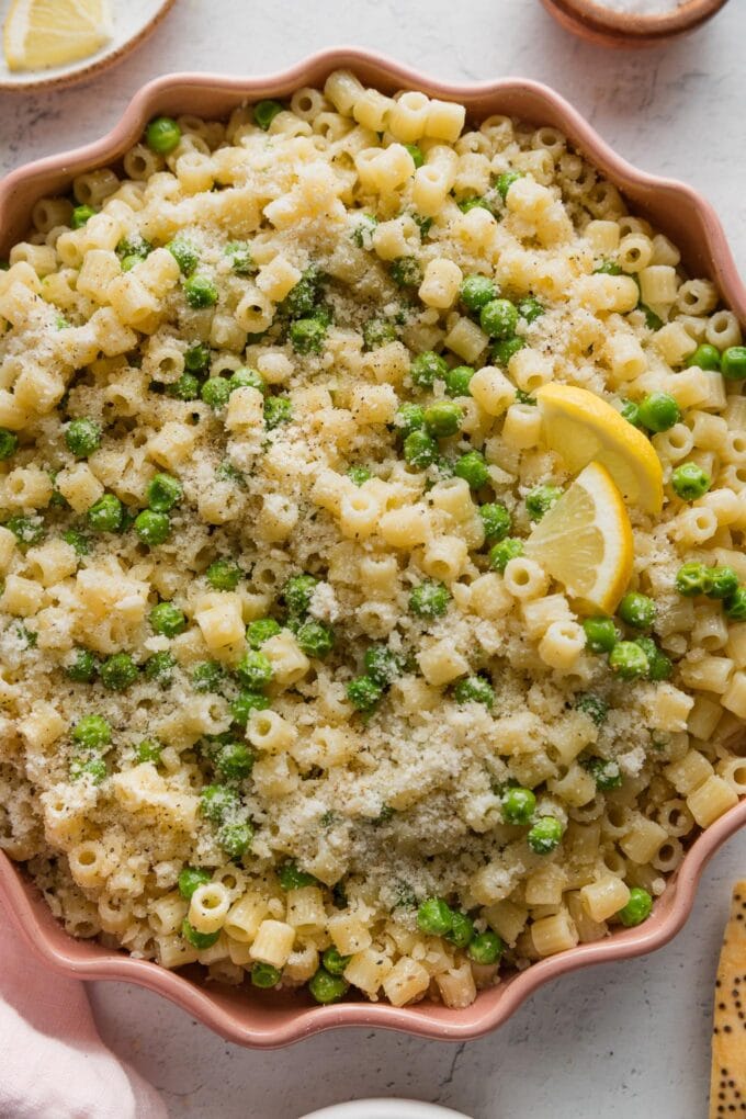 Close up of ditalini with peas, Parmesan, and black pepper served in a pink scalloped bowl with extra Parmesan on the table nearby.