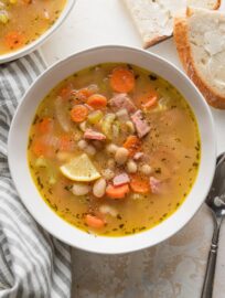 Bowl of ham and white bean soup garnished with a lemon wedge and with fresh buttered bread on the counter nearby.