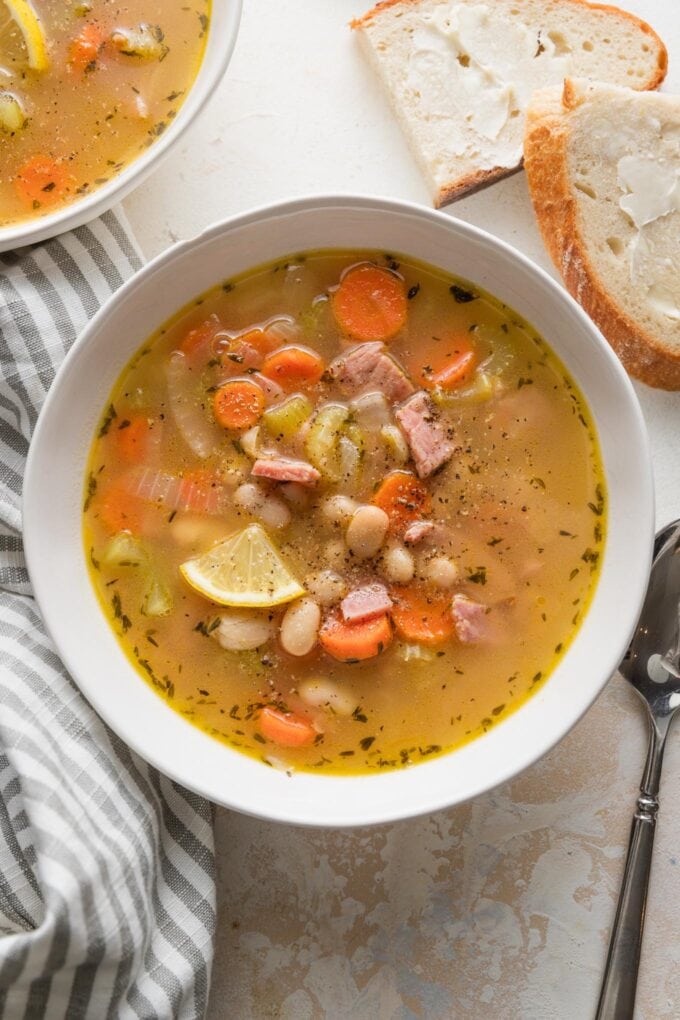 Bowl of ham and white bean soup garnished with a lemon wedge and with fresh buttered bread on the counter nearby.
