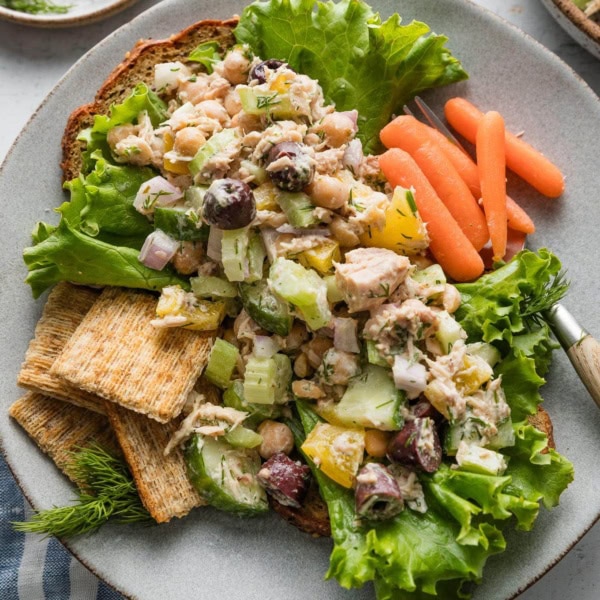 Plate full of a Mediterranean-inspired tuna salad served over toast and lettuce with baby carrots and extra olives on the side.