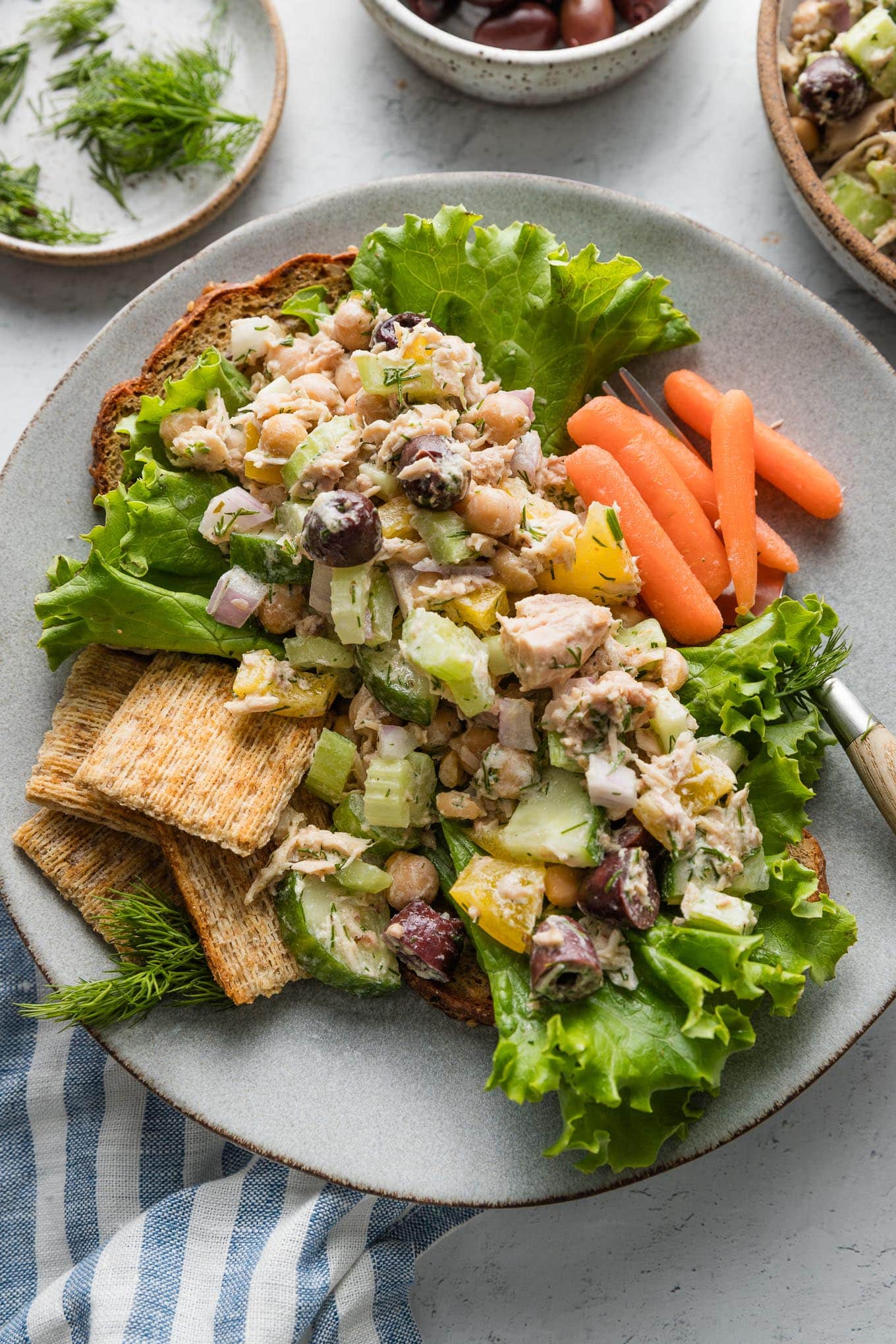 Plate full of a Mediterranean-inspired tuna salad served over toast and lettuce with baby carrots and extra olives on the side.