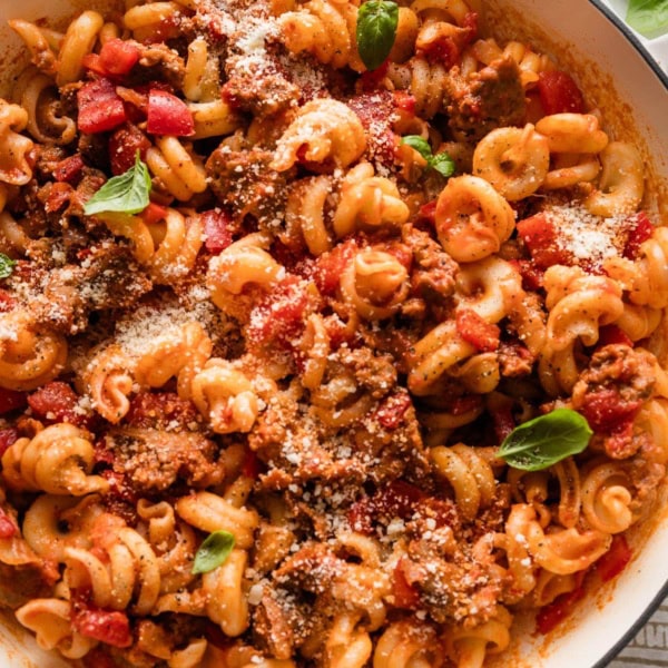 Close up of sausage and peppers pasta in a large skillet garnished with Parmesan and fresh basil leaves.