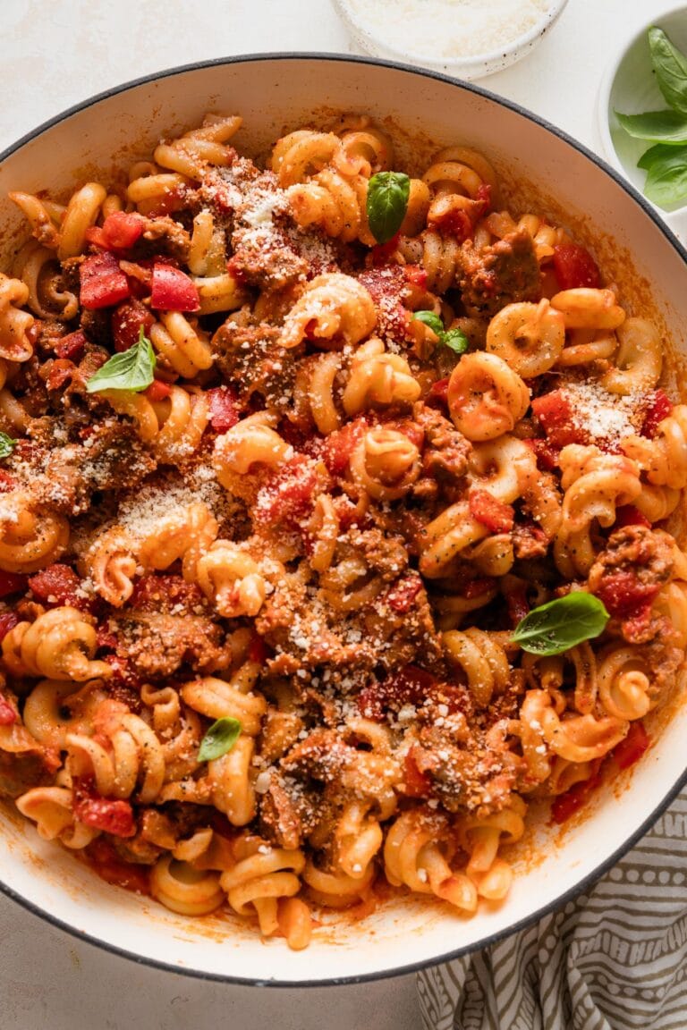 Close up of sausage and peppers pasta in a large skillet garnished with Parmesan and fresh basil leaves.