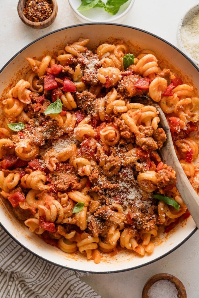 Wooden spoon lifting a helping of sausage pepper pasta out of a large skillet to serve.