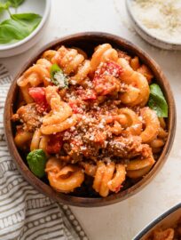Small wooden bowl with a helping of sausage and peppers pasta topped with fresh Parm and basil.