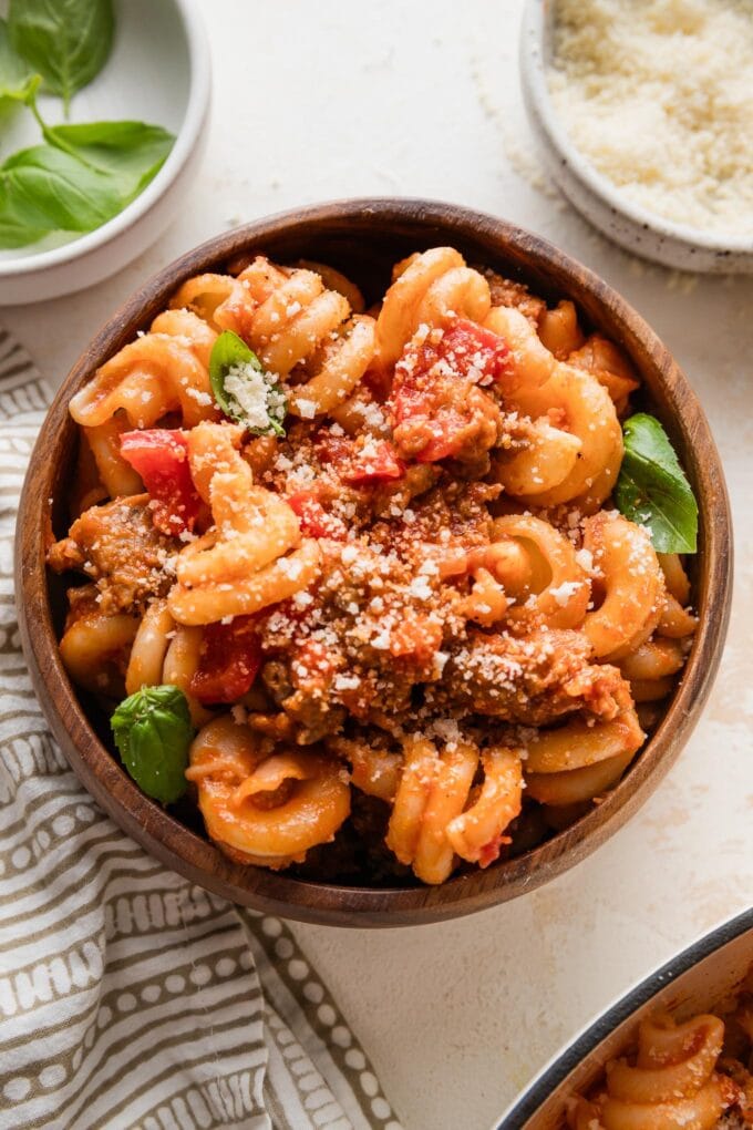 Small wooden bowl with a helping of sausage and peppers pasta topped with fresh Parm and basil.