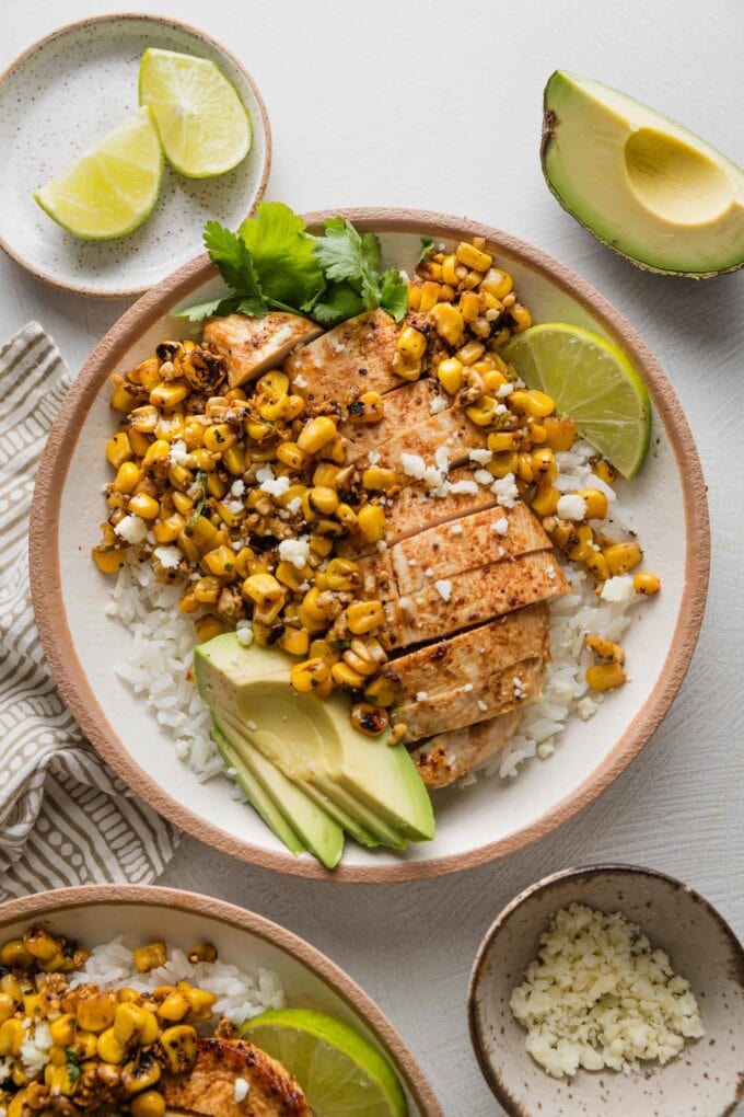 Countertop set with two street corn chicken and rice bowls, with extra lime wedges and avocado nearby.