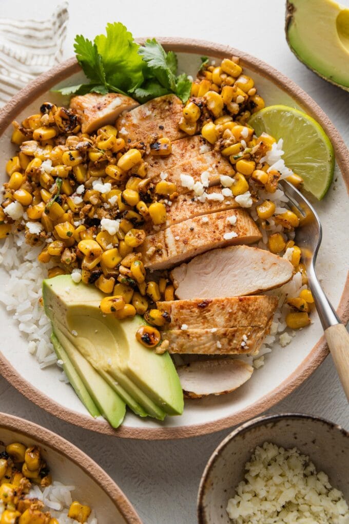 Close up of a rice bowl with Tajin-seasoned chicken breast, street corn with Cotija, avocado, fresh cilantro, and lime.