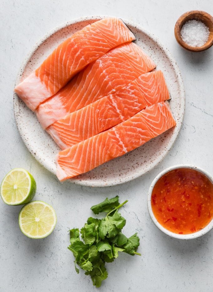 Four salmon filets on a plate, with Thai sweet chili sauce, a fresh lime, kosher salt, and cilantro leaves scattered nearby.