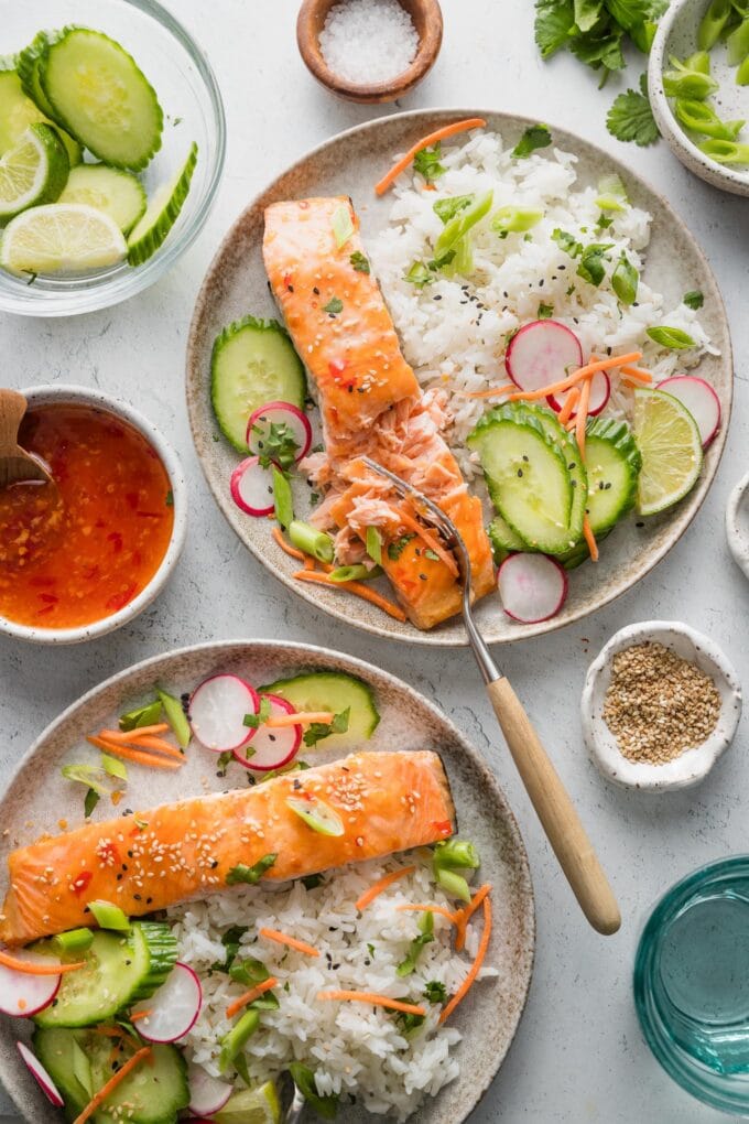 Countertop set with two dinner plates of Thai-inspired sweet chili salmon with rice, cucumber, radish, shredded carrot, cilantro, and lime.