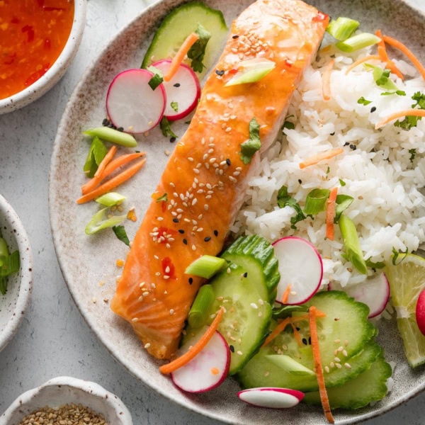Plate of sweet chili lime salmon served with rice, cucumber, radish, cilantro, lime, and carrots.