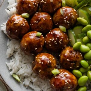 Close up of Asian-inspired chicken meatballs in a sweet and sticky homemade glaze plated over rice with cucumber and edamame.