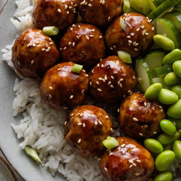 Close up of Asian-inspired chicken meatballs in a sweet and sticky homemade glaze plated over rice with cucumber and edamame.