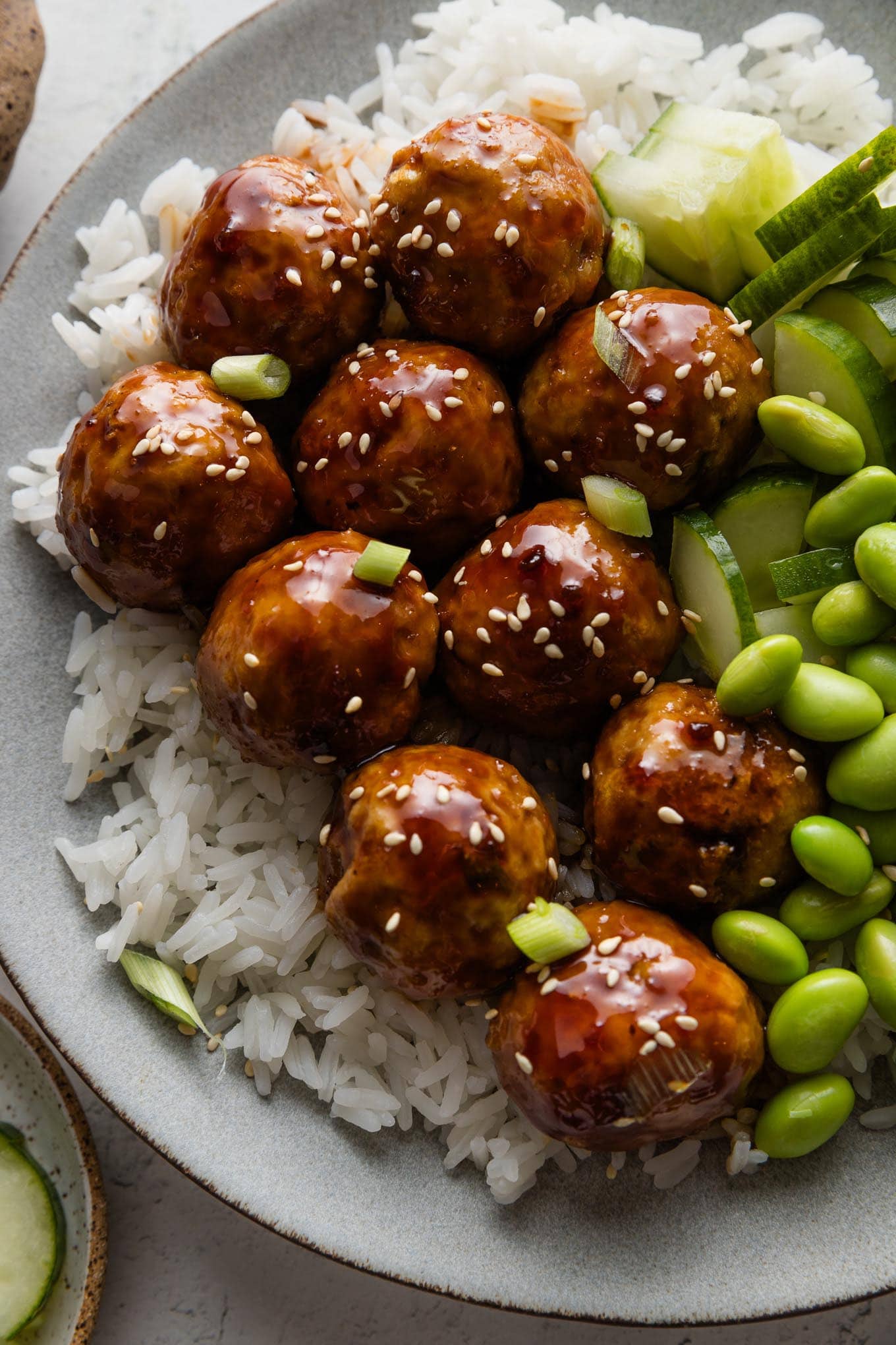 Close up of Asian-inspired chicken meatballs in a sweet and sticky homemade glaze plated over rice with cucumber and edamame.