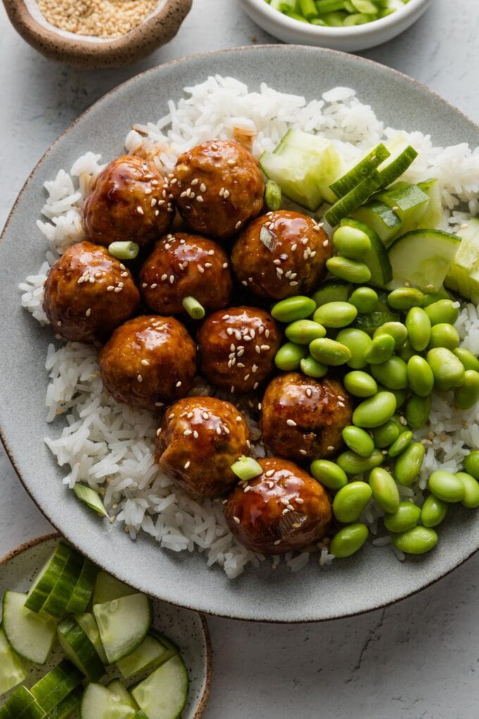 Plate of Asian chicken meatballs over rice with edamame, cucumber, homemade glaze, sesame seeds, and green onions.