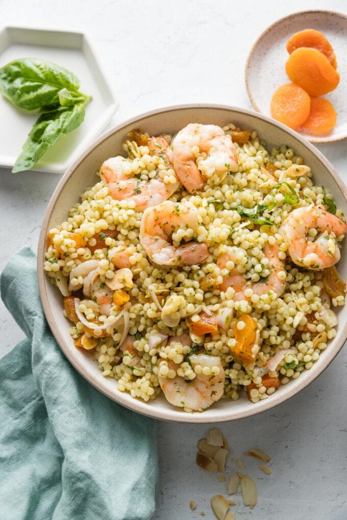 Countertop set with a bowl of pearl couscous salad with poached shrimp, dried apricots, and basil vinaigrette, with extra basil leaves and dried apricots in small bowls nearby.