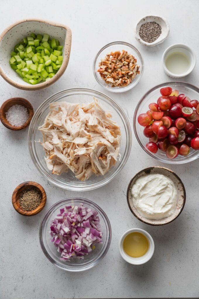 Flatlay with bowls of shredded chicken, red grapes, chopped celery and red onion, Greek yogurt, apple cider vinegar, honey, salt, and pepper.