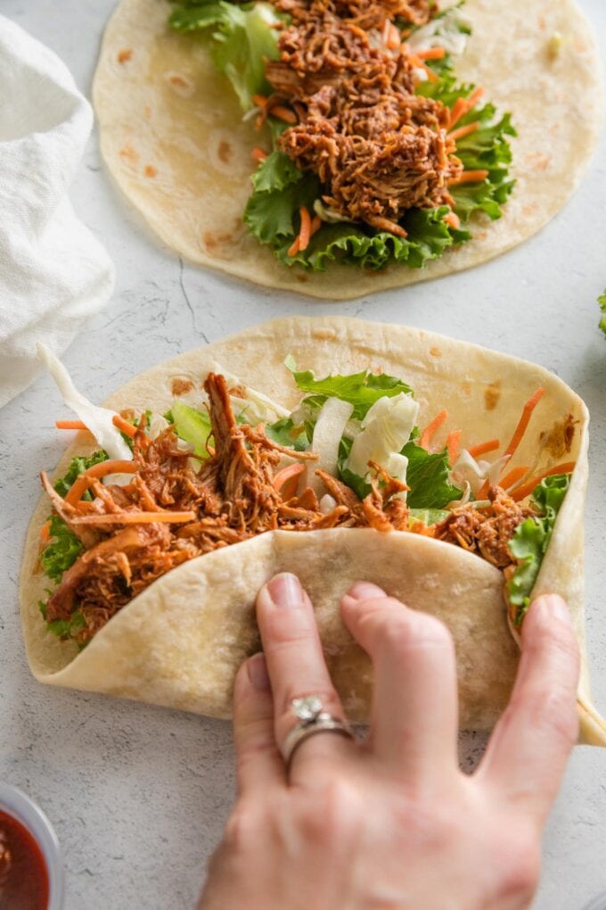 Woman's hand folding up a tortilla around lettuce, veggies, and BBQ chicken.