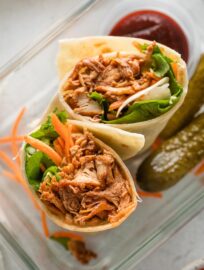 BBQ chicken wrap cut in half and tucked into a glass container along with extra BBQ sauce and two small dill pickles.