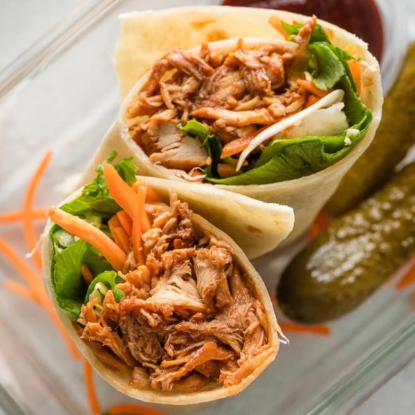 BBQ chicken wrap cut in half and tucked into a glass container along with extra BBQ sauce and two small dill pickles.