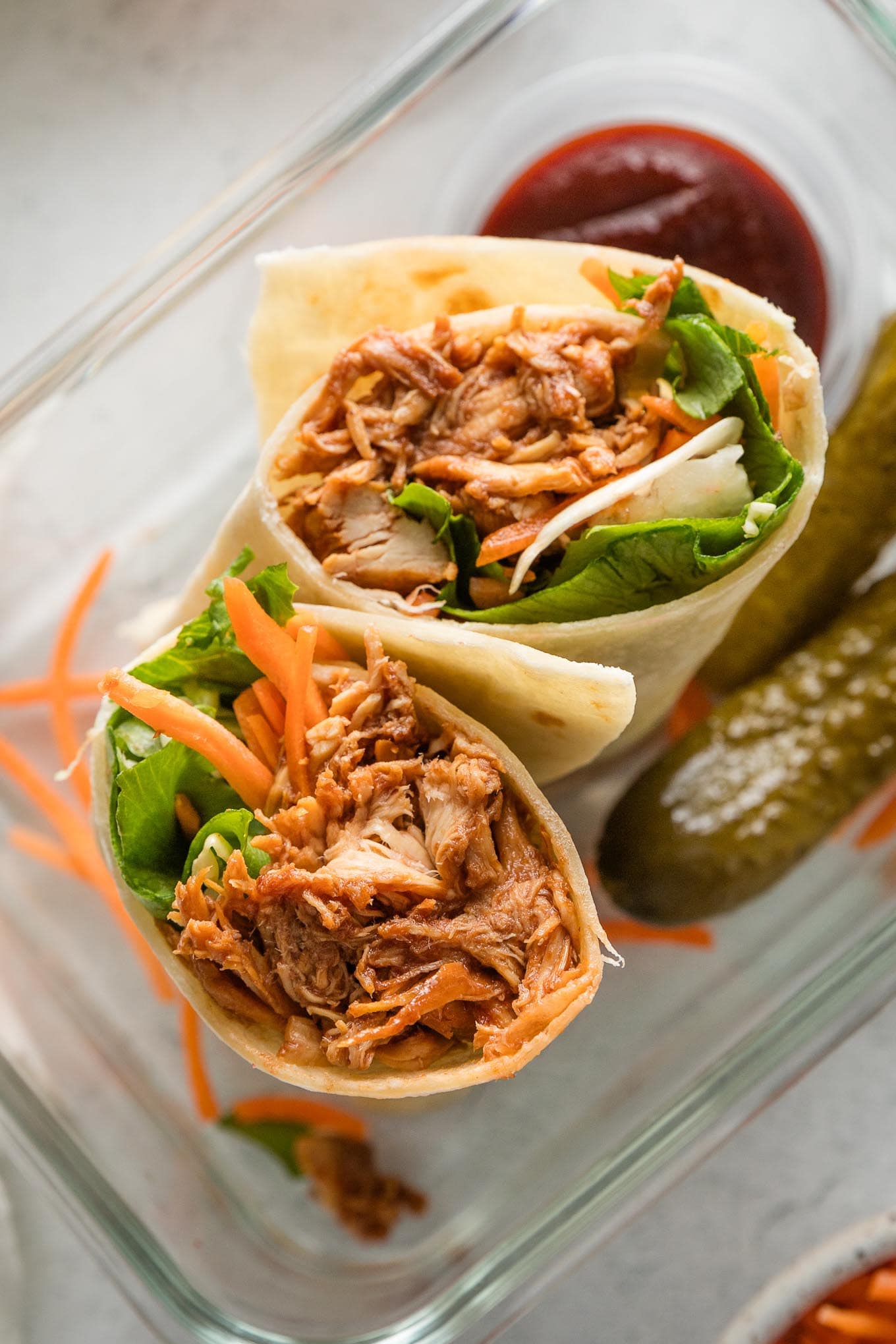 BBQ chicken wrap cut in half and tucked into a glass container along with extra BBQ sauce and two small dill pickles.