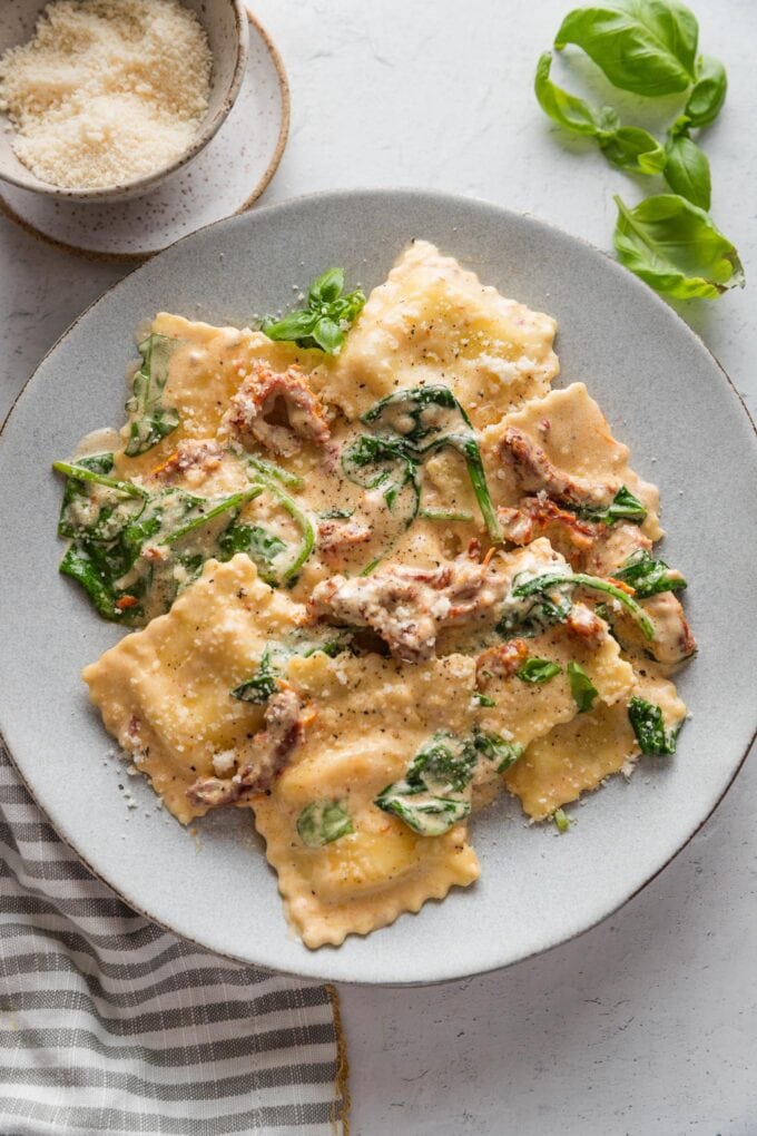 Overhead view of a small dinner plate full of creamy Tuscan ravioli with spinach and sun-dried tomatoes, with extra grated Parmesan and basil leaves scattered nearby on the counter.