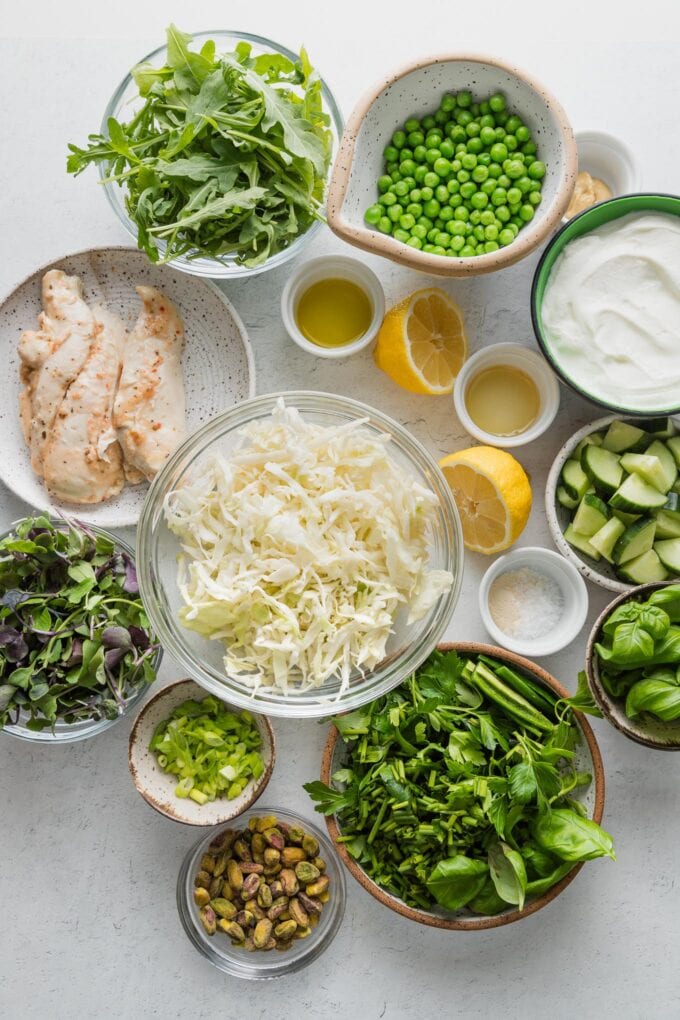 Flatlay of ingredients for a green goddess salad--green cabbage, microgreens, arugula, basil, peas, pistachios, Greek yogurt, lemon, and more--in small prep bowls.