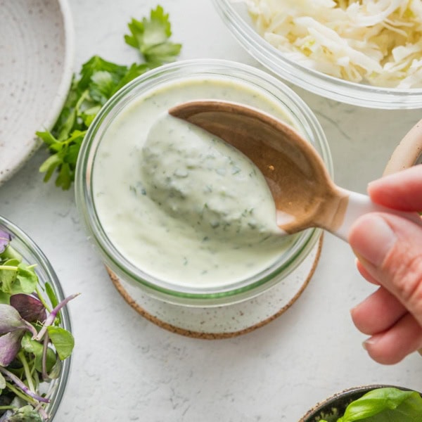 Close-up of homemade green goddess dressing in a small glass jar being lifted out with a small ceramic spoon.