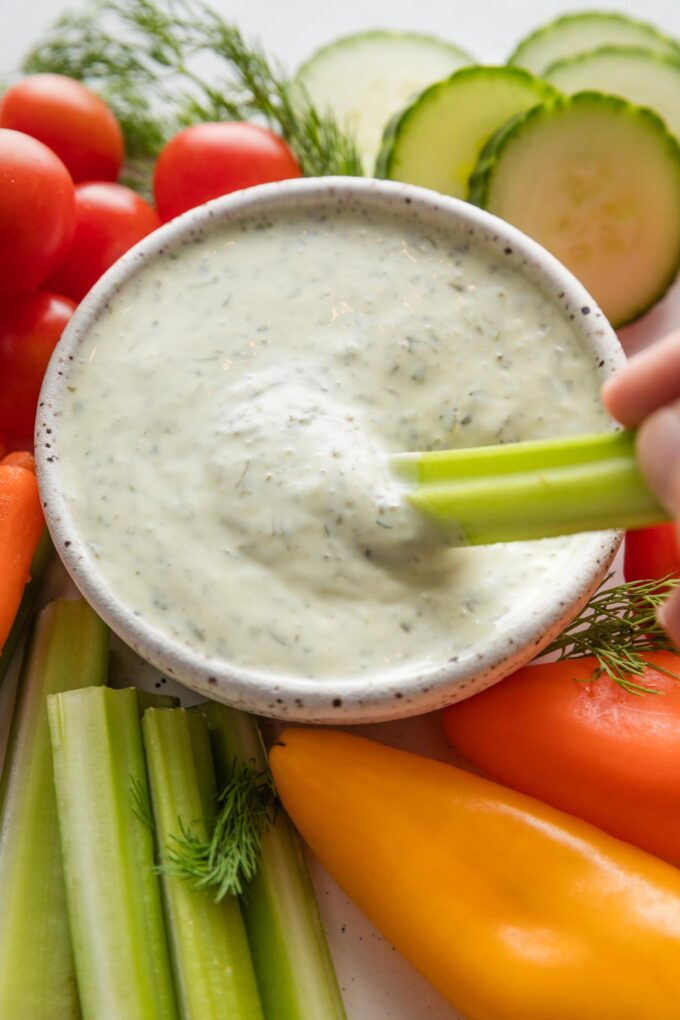 Close up of a woman's hand dipping a celery stick into a bowl of homemade green goddess dressing.