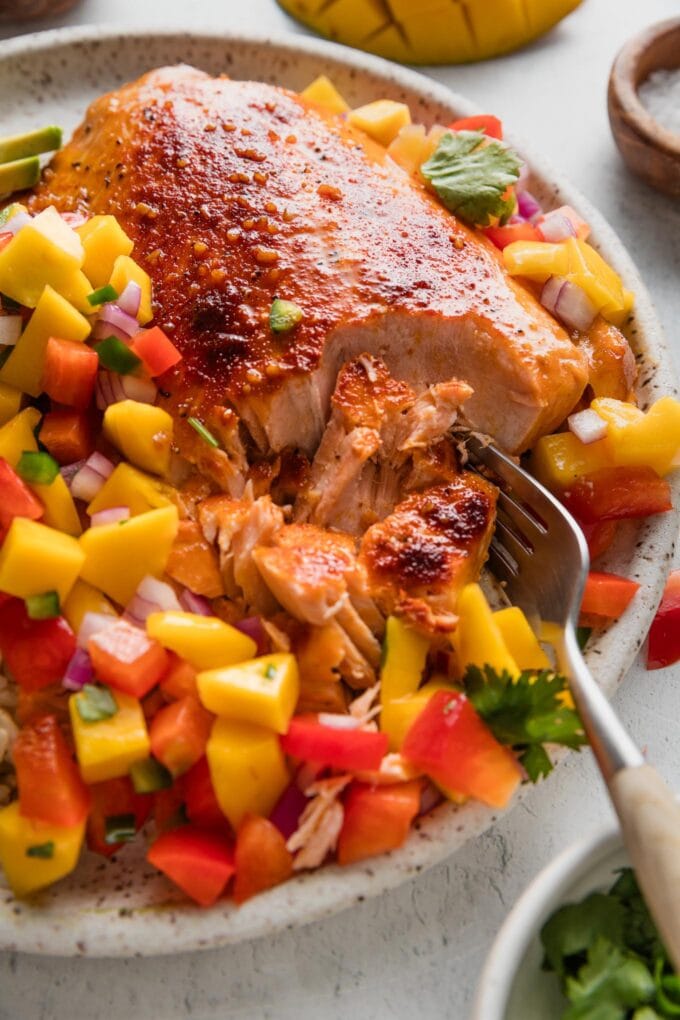 Close up of a fork flaking off a piece of honey lime seasoned salmon on a plate with mango salsa.