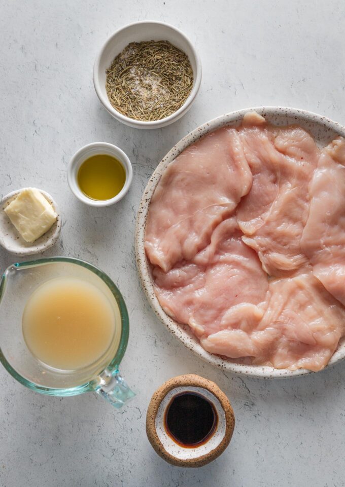 Flatlay photo of thin-sliced chicken breasts, balsamic vinegar, herbs, butter, garlic, and chicken broth.