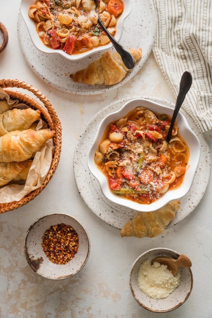 Bowls of minestrone soup with chicken set on a counter to serve alongside extra Parmesan, red pepper flakes, and mini croissants.