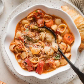 Small white scalloped bowl full of chicken minestrone soup with sun-dried tomato pesto, extra Parmesan, and red pepper flakes.