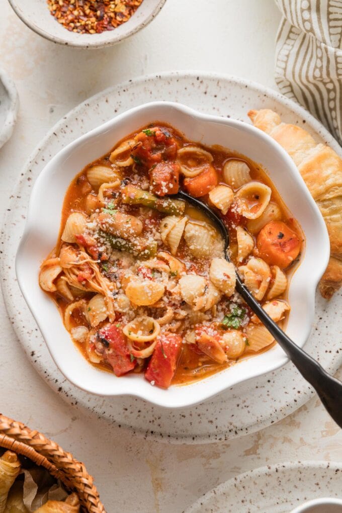 Small white scalloped bowl full of chicken minestrone soup with sun-dried tomato pesto, extra Parmesan, and red pepper flakes.
