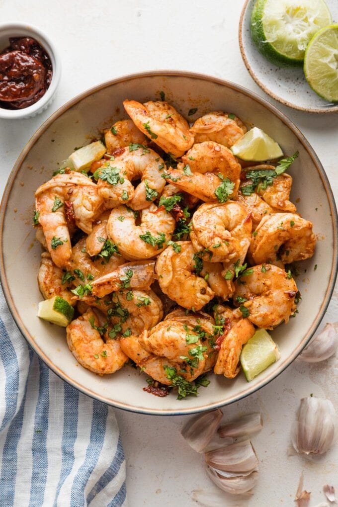Bowl full of oven-baked chipotle shrimp resting on a counter with extra garlic cloves, lime, and adobo sauce on the counter nearby.