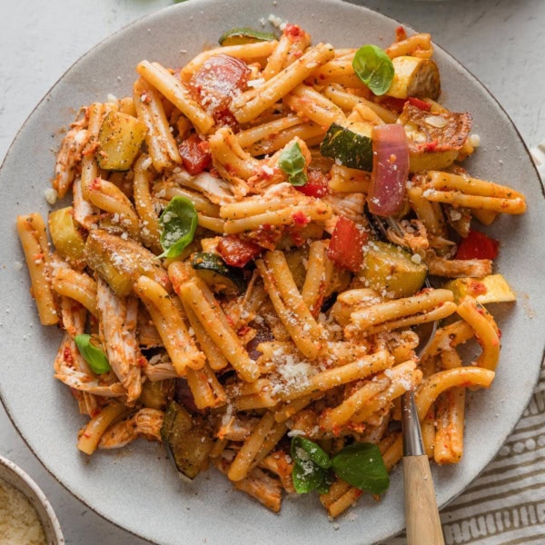 Plate full of pasta with a light sun-dried tomato pesto sauce, shredded rotisserie chicken, and roasted balsamic veggies, all mixed together and topped with fresh basil and Parmesan.