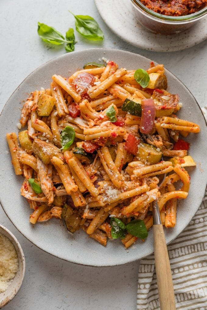 Plate full of pasta with a light sun-dried tomato pesto sauce, shredded rotisserie chicken, and roasted balsamic veggies, all mixed together and topped with fresh basil and Parmesan.