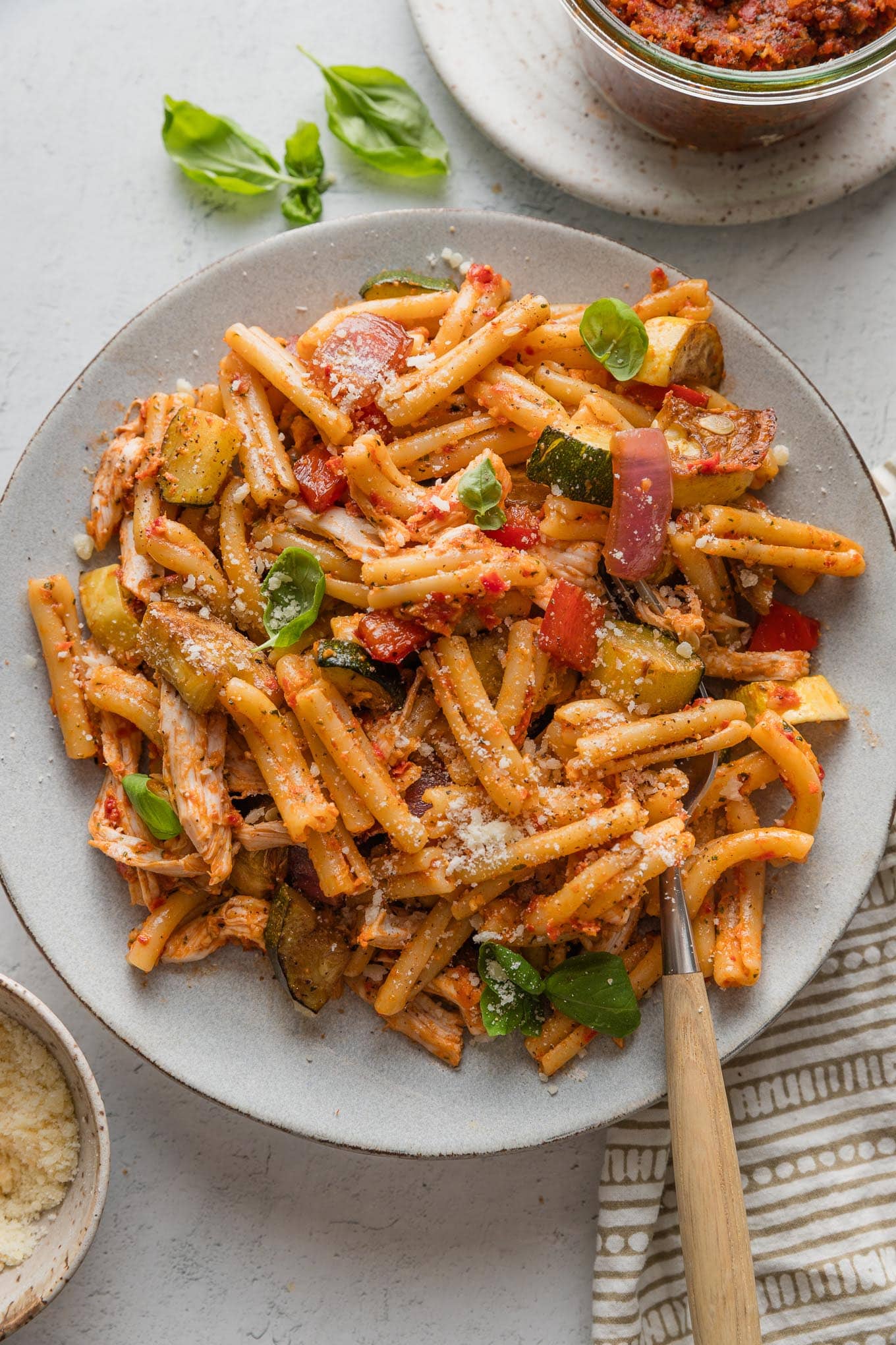 Plate full of pasta with a light sun-dried tomato pesto sauce, shredded rotisserie chicken, and roasted balsamic veggies, all mixed together and topped with fresh basil and Parmesan.