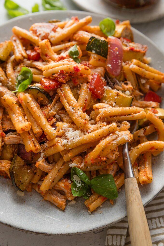 Close up of a plate filled with sun-dried tomato pesto pasta with shredded rotisserie chicken and roasted veggies mixed in.