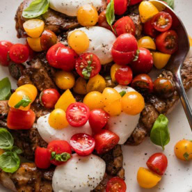 Spoon lifting a generous helping of cherry tomatoes over a platter of balsamic-marinated chicken breasts.