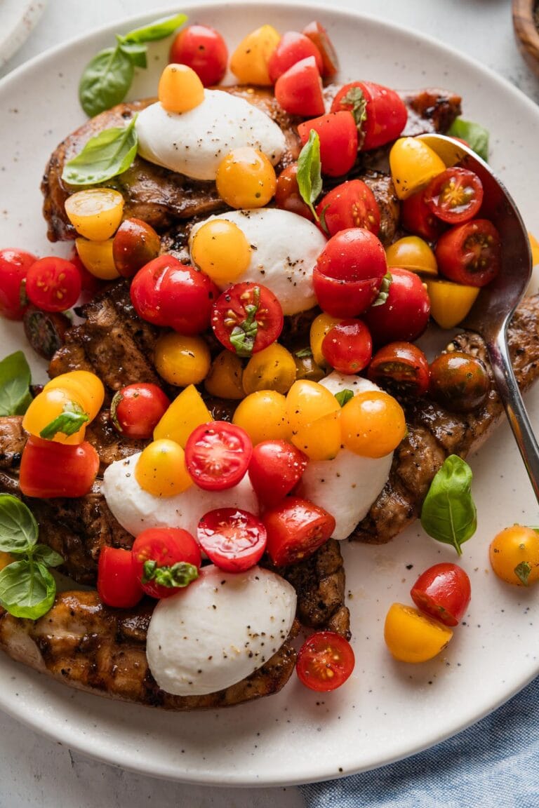 Spoon lifting a generous helping of cherry tomatoes over a platter of balsamic-marinated chicken breasts.