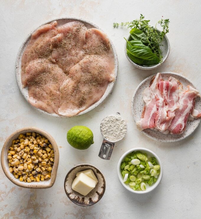 Overhead view of thin-sliced chicken breasts, strips of bacon, corn kernels, butter, olive oil, all-purpose flour, a lime, green onions, and fresh herbs.