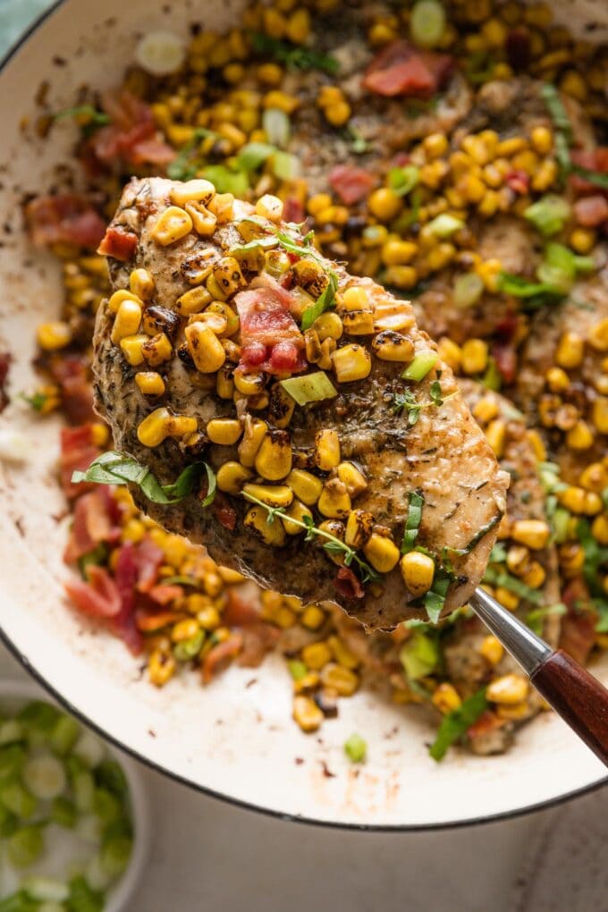 Spatula lifting a serving of chicken with brown butter corn and bacon out of a deep skillet.
