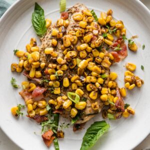Plate with a helping of chicken breast pan-fried and topped with brown butter corn, bacon, and fresh herbs and green onions.